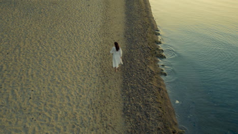 rearview tracking follows beautiful woman in white flowy airy dress on beach by ocean