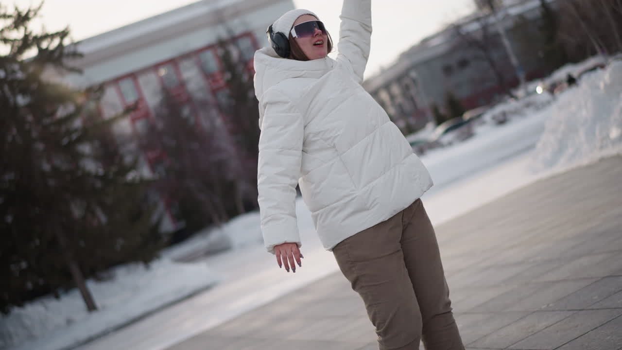 Closeup of young woman wearing fluffy white jacket, beanie, sunglasses and headphones, throwing hands up in air and moving body with excitement on snow dusted urban plaza under pale winter light