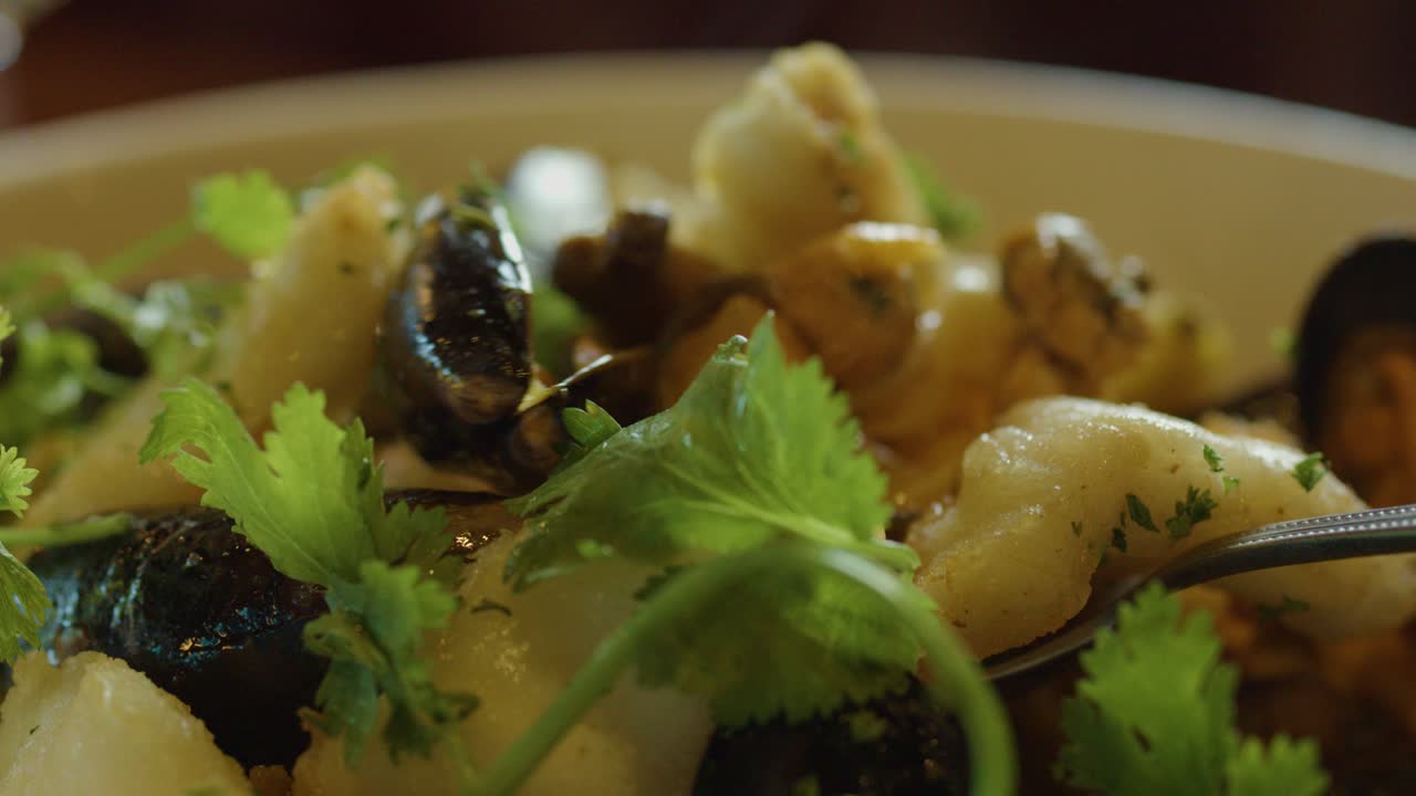 A close-up view of a hand using a spoon to serve hot seafood chowder with mussels, squid, scallops, and fresh cilantro in warm, natural lighting