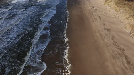 Aerial-view-captures-the-beauty-of-a-beach,-with-cloud-shadows-dancing-over-the-sand-dunes,-waves-crashing-onto-the-shore,-birds-soaring-overhead,-and-a-lone-figure-strolling-along-the-beach