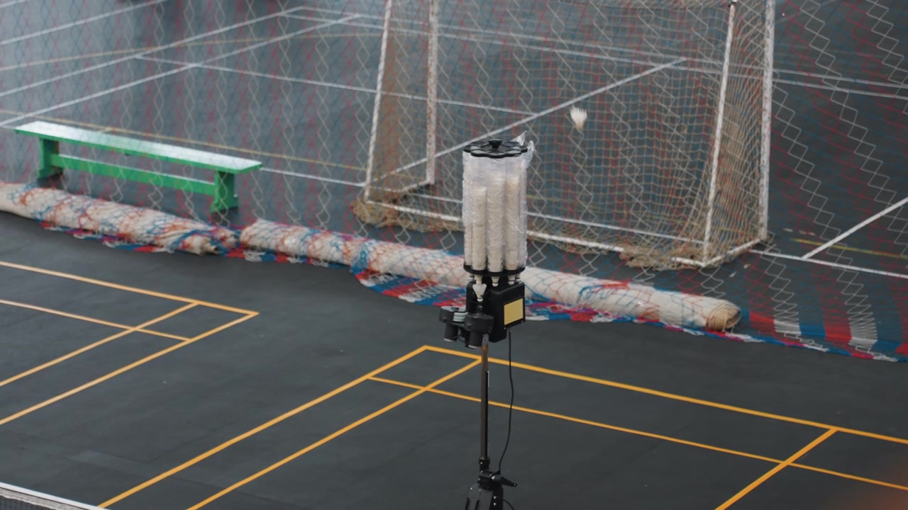 Empty indoor badminton court with launcher shuttlecock machine on tripod near net green bench along sideline yellow court lines colorful barrier behind net single shuttle on floor quiet gym atmosphere