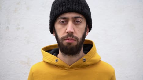 Bearded Man With Serious Face In Mustard Hoodie And Knitted Beanie Pointing Finger At His Eye Showing To Someone I Am Watching You Standing Against A Concrete Wall