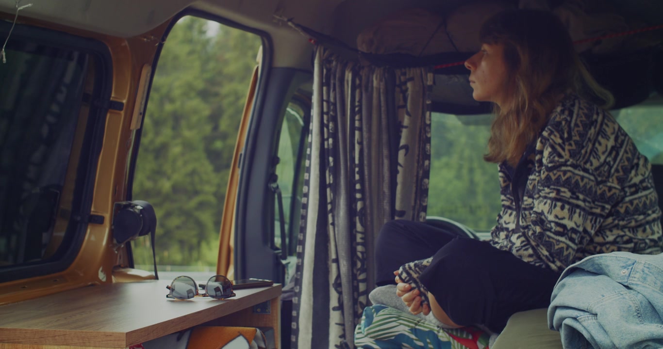 Una giovane donna siede comodamente all'interno del suo camper, guardando pensierosa fuori dalla finestra verso una lussureggiante foresta verde, incarnando un senso di libertà e voglia di viaggiare.