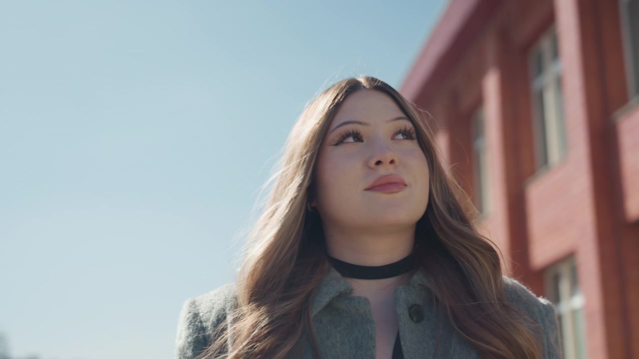Donna asiatica riflessiva. Primo piano di donna contemplativa in piedi davanti a un edificio in mattoni che sorride con sicurezza. Vista ravvicinata di donna asiatica introspettiva che indossa un cappotto contro un muro di mattoni in una luminosa giornata di sole.