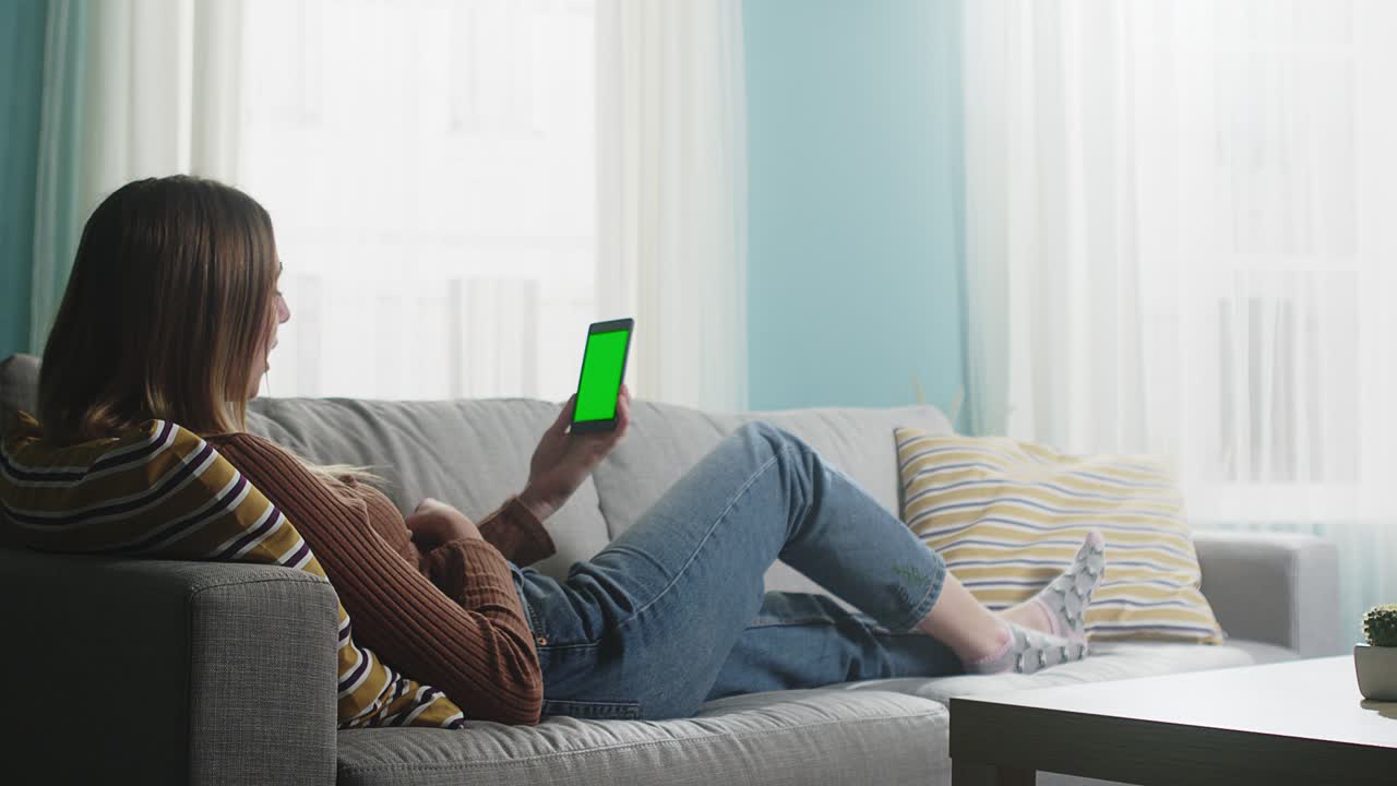 젊은 여자가 베이지 소파에 누워서 비디오 통화를 하고 있습니다.