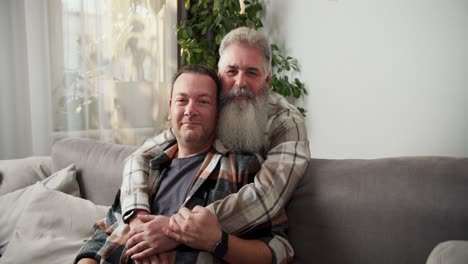 A-happy-elderly-man-with-gray-hair-and-a-lush-and-gray-beard-in-a-checkered-shirt-hugs-his-beloved-brunette-boyfriend-in-a-checkered-shirt-while-sitting-on-a-gray-sofa-in-a-modern-apartment