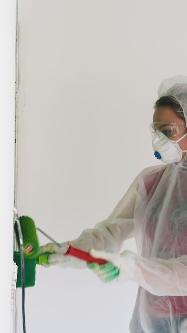 young man and woman in transparent coveralls and respirators paint white wall with green color using rollers in light room