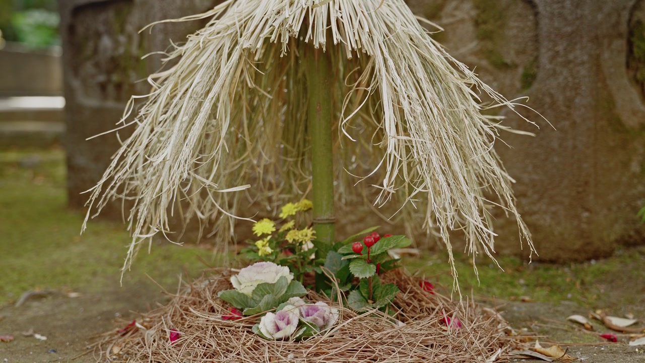 Tilt-down shot revealing a traditional straw canopy decoration above a woven nest base with rose and yellow flower offerings at the base of a sacred shrine tree in natural daylight.