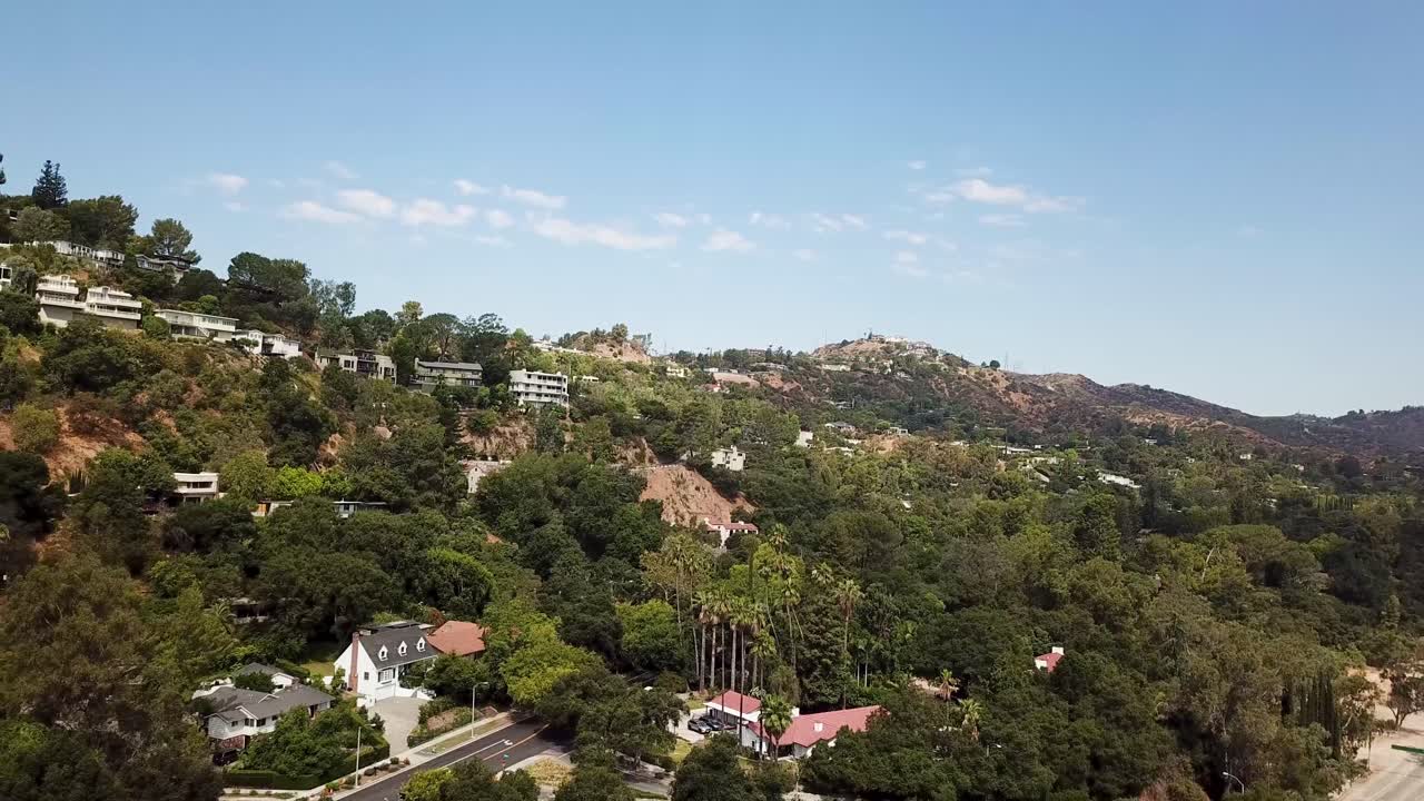 Aerial view of hillside homes in California surrounded by lush greenery and palm trees. Scenic residential architecture with mountain backdrop under clear blue sky. Rising shot. Pasadena, USA