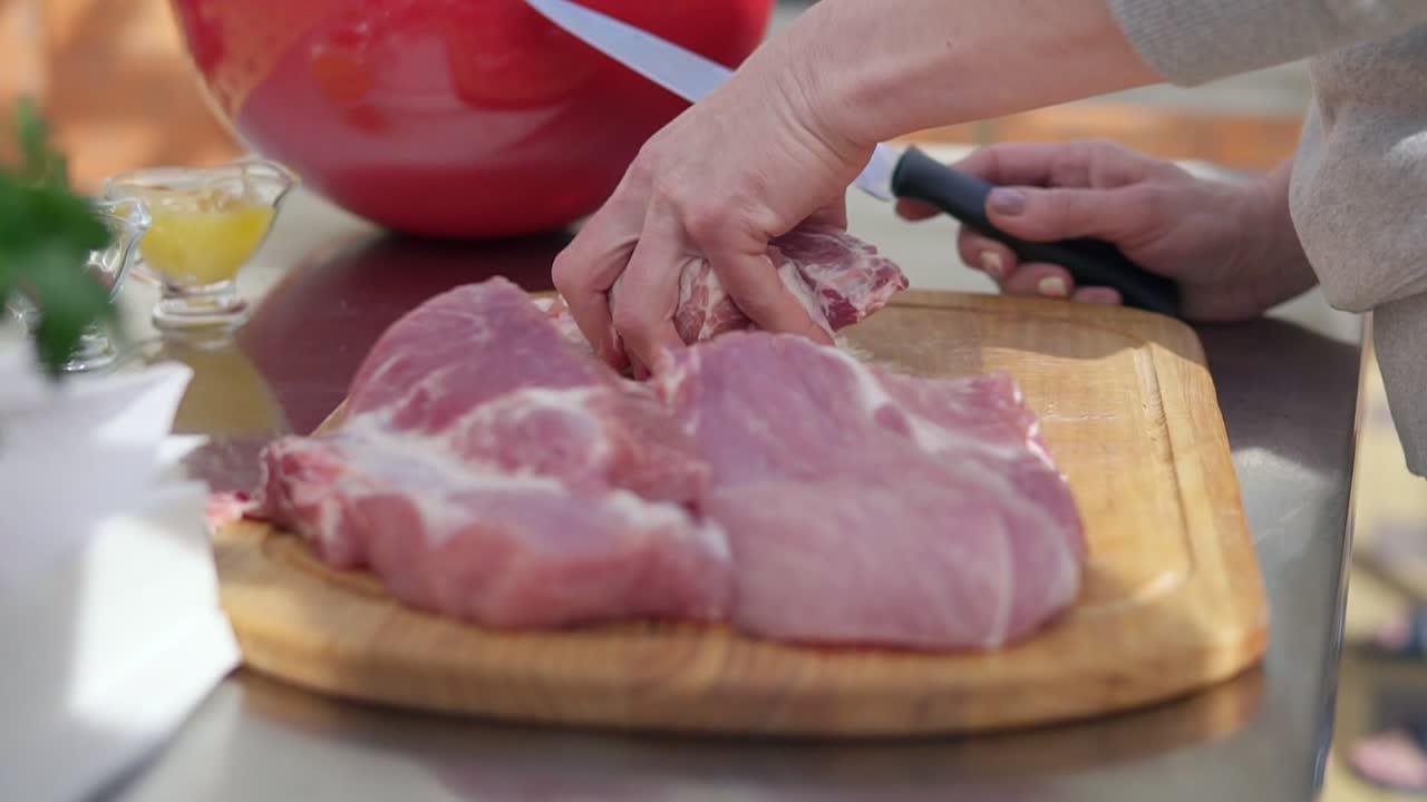 Person's hands cutting into a large piece of raw meat using a butcher's knife on a wooden surface outside. Barbeque preparation