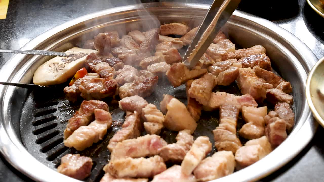 Tongs turn sizzling pork belly pieces on tabletop grill under bright lighting, close-up overhead view