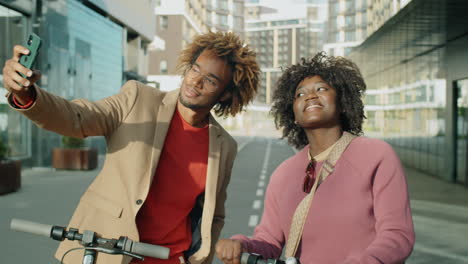 joyous african american friends taking selfie with e-scooters in city