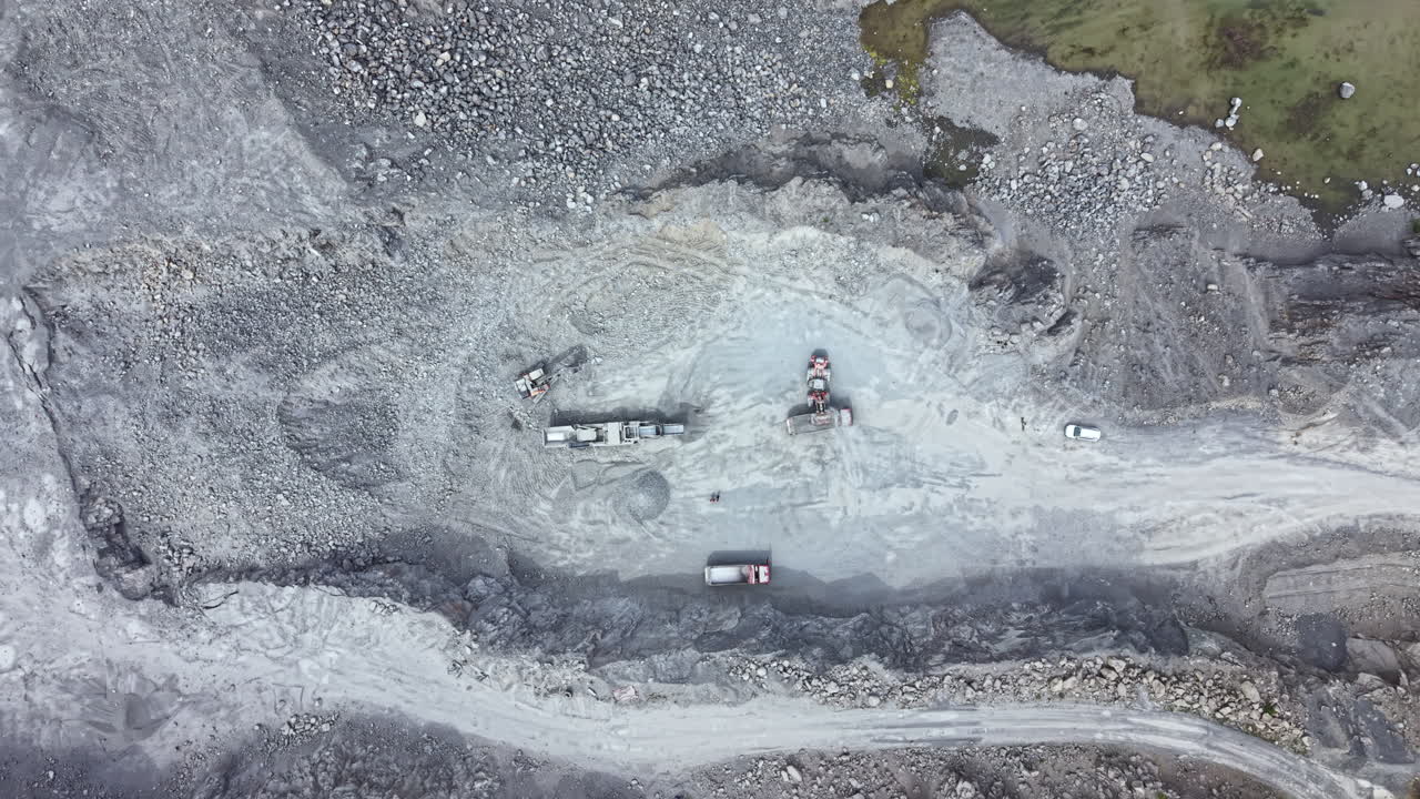 Construction activity takes place at a quarry site with heavy machinery working on rock mining and material transport during daylight hours in an open landscape