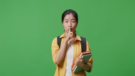 녹색 스크린 배경 스튜디오에 서있는 동안 카메라를보고 shh 제스처를하는 배과 몇 권의 책을 가진 아시아 여성 학생