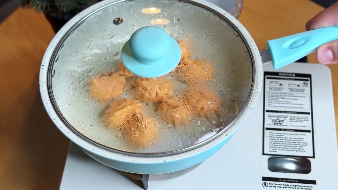 Primo piano di una mano che posiziona una padella blu coperta su un fornello portatile bianco. La condensa si forma sul coperchio di vetro mentre alimenti arancioni cuociono a vapore all'interno su un caldo tavolo da cucina in legno