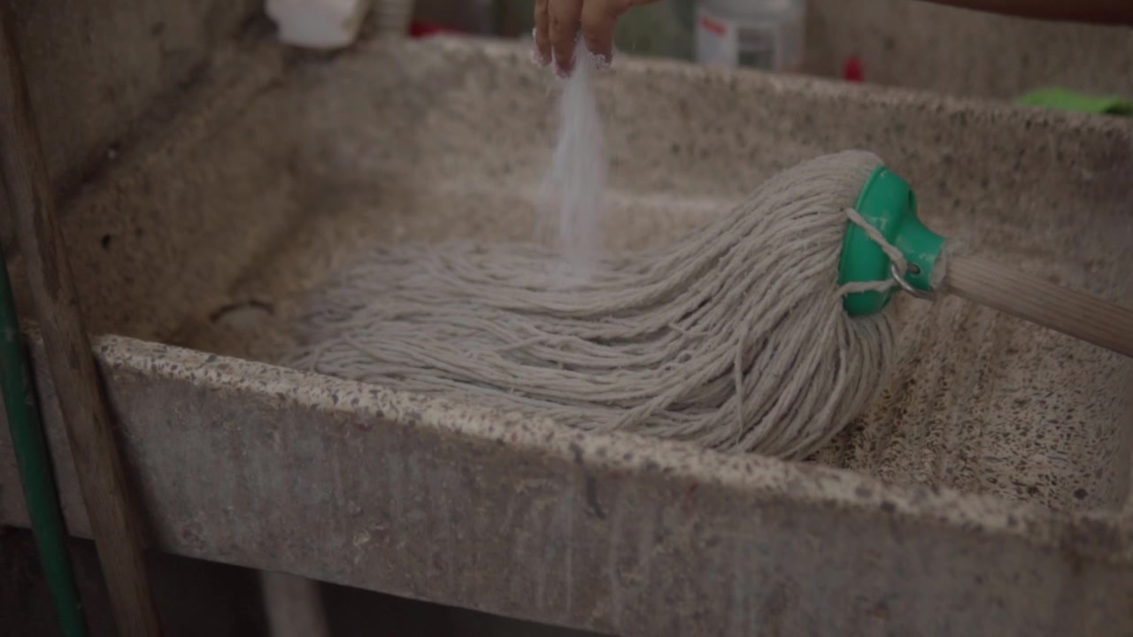 Versare detersivo in polvere sul mocio e lavare con acqua in un lavatoio, mostrando il processo di pulizia, fibre bagnate, residui di sapone e lavabo in cemento