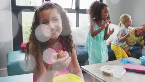 animation of white bokeh light spots over happy diverse schoolgirls eating packed lunch and talking