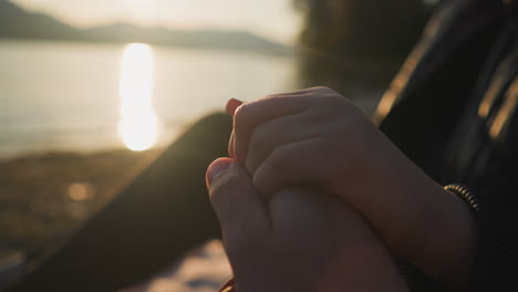 lovers hold hands sitting at sunset closeup. boyfriend enjoys romantic atmosphere of date with girlfriend on riverbank together. heartwarming moment