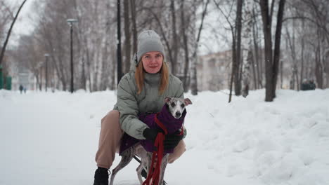 dog owner kneeling on snowy forest path holding whippet dressed in purple coat, both facing forward, woman wearing green winter jacket and gray beanie, surrounded by snow-covered ground and trees