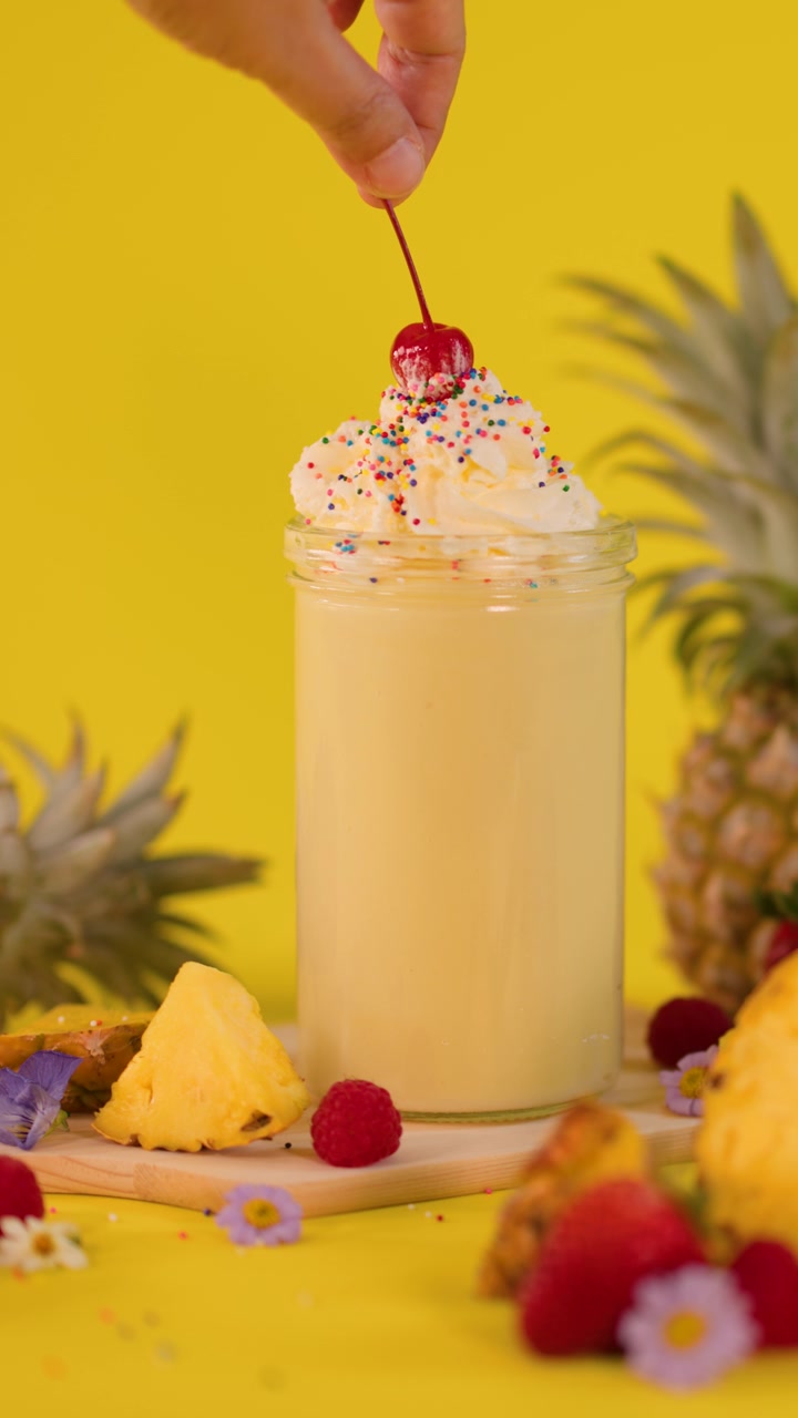 Una mano posiziona una ciliegia sulla panna montata di un milkshake all'ananas, circondato da frutta fresca, con illuminazione brillante.