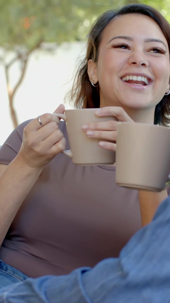 Video vertical: Mujer bebiendo de una taza beige en un sofá exterior, brindando, charlando y riendo.