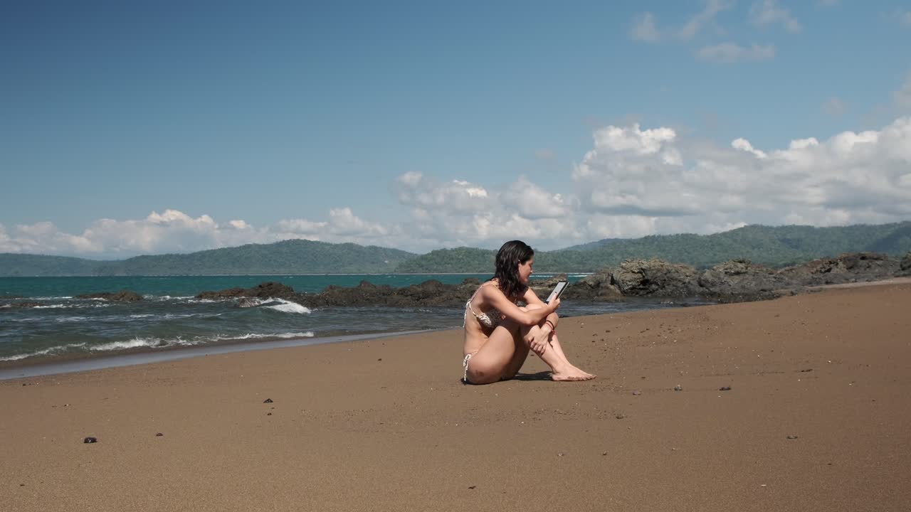 mujer leyendo un libro electrónico en una serena playa tropical junto a la orilla del océano