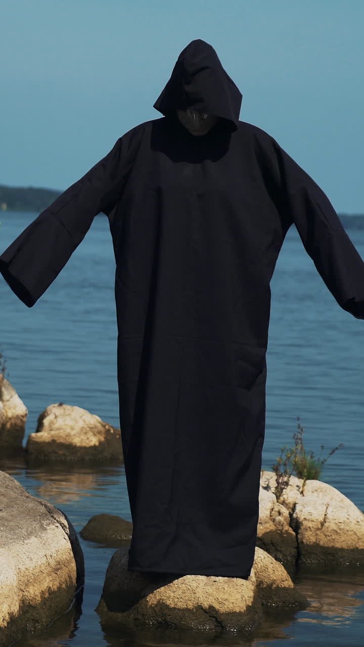Woman devil in black cape and hood on stones near the river. Halloween costume.. Vertical video