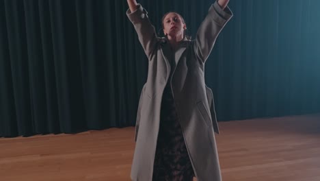 woman in coat performs emotional solo dance on wooden floor under stage lights