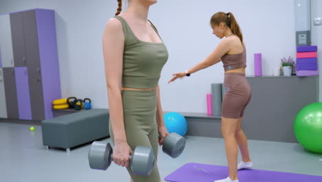 woman in sports outfit lifts dumbbells from purple mat during gym workout while sisters call out to someone in background, engaging in training session