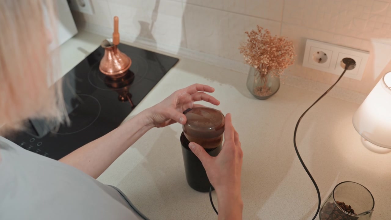 Femme blanche moulant des grains de café sur le comptoir, sous la lueur d'une lampe, avec une bouilloire en cuivre et des fleurs séchées encadrant la scène, un moulin électrique ronronnant, une cuillère mesurant le café moulu, les mains stables, dans une cuisine minimaliste et confortable, intime.