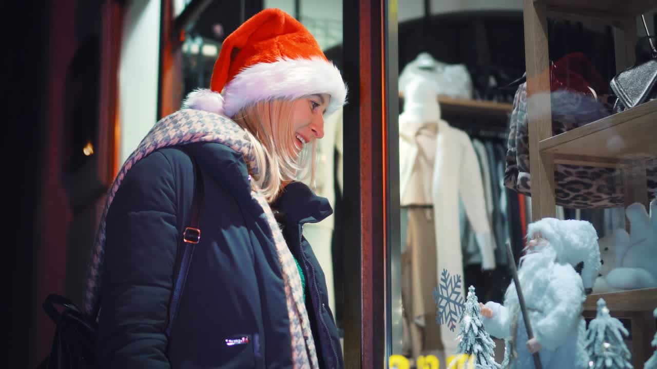 산타 모자를 입은 여자가 밤에 가게 창문을 바라보고 있다.