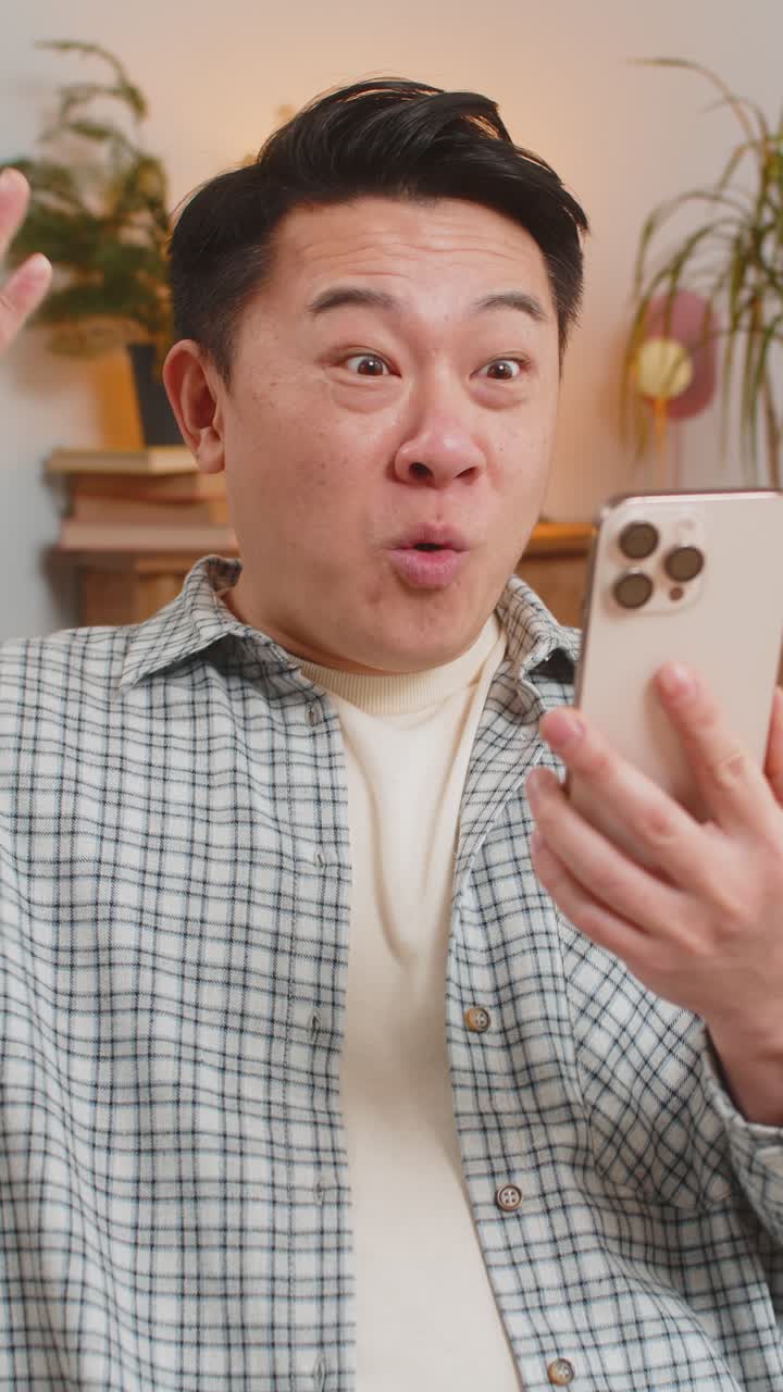 Uomo asiatico di mezza età a casa che celebra la vittoria di un gioco online scioccato da buone notizie al telefono