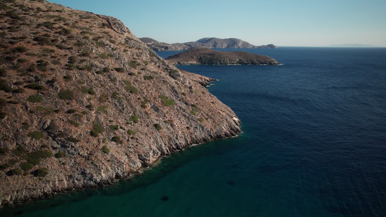 Carrellata aerea lungo la costa frastagliata, sabbiosa ed erosa di Syros, Grecia, per rivelare un villaggio distante, a mezzogiorno.