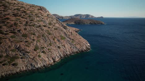 Aerial-dolly-along-rugged-sandy-eroded-coastline-of-syros-greece-to-reveal-distant-village,-midday