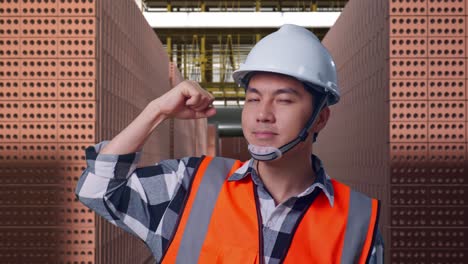 Close Up Of Asian Male Engineer With Safety Helmet Flexing His Bicep And Smiling To Camera While Standing With Red Brick Packed in Stacks Are Stored