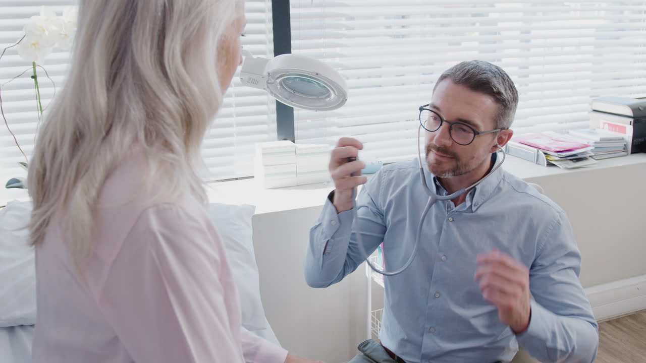médico escuchando a una paciente femenina madura respirando con estetoscopio durante un examen médico en la oficina