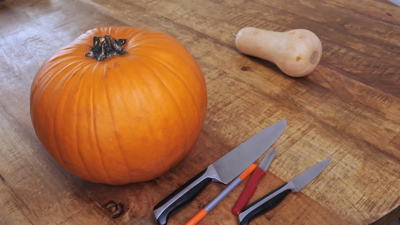 A man prepares to cut a fresh pumpkin for Halloween decoration on a wooden table with sharp knives