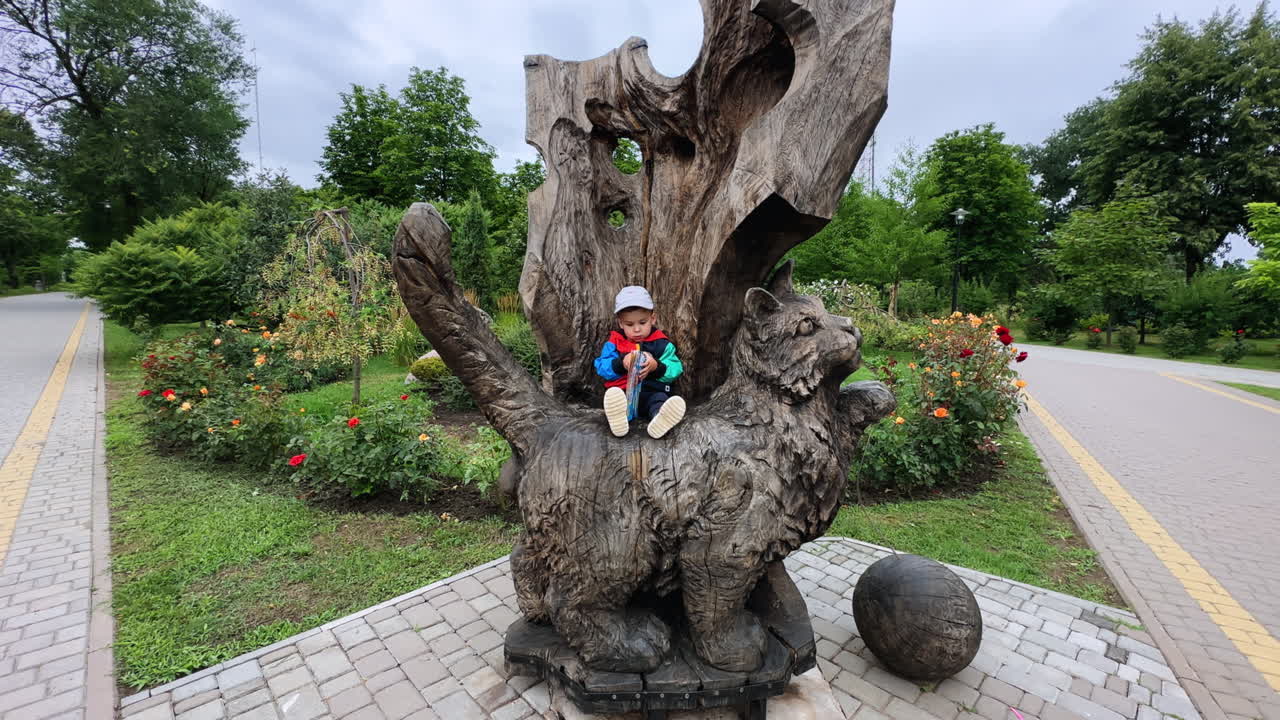 Small Caucasian toddler sits on a big wooden cat in the park. Lovely kid looks through the book.
