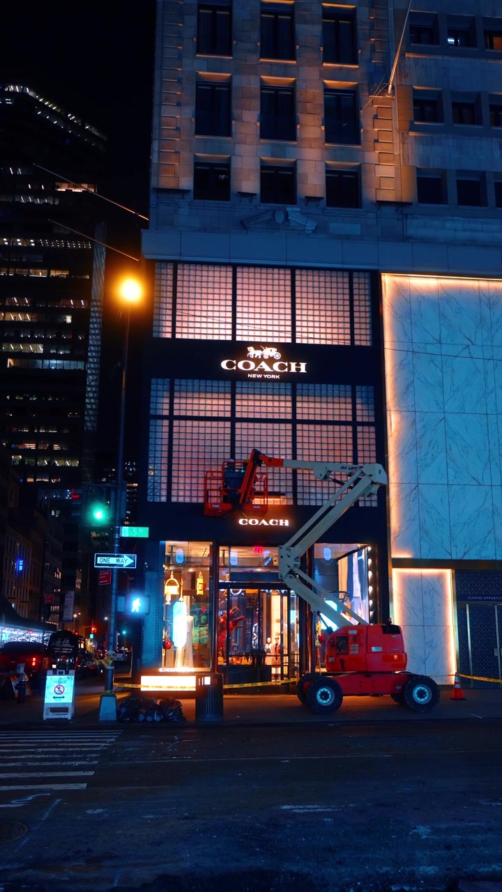 New York, USA, 15 aprile 2026: Piattaforma aerea fuori dal negozio Coach sulla Fifth Avenue. Una piattaforma aerea arancione è parcheggiata di fronte alla vetrina di Coach New York sulla Fifth Avenue di notte.