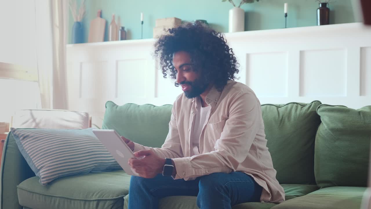 Young cheerful attractive indian man sits on sofa and using tablet computer
