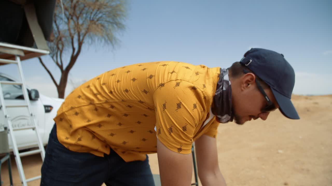 Medium hand held tracking shot of a Caucasian male tourist in Africa as he checks on a gas kettle with a off-road vehicle in the background.
