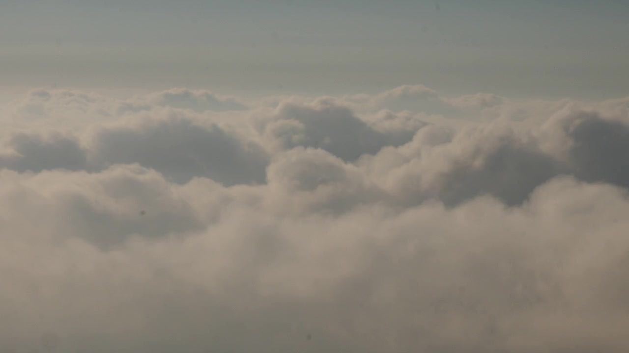 Clouds drift across a pale blue sky in a wide aerial view above a dense cloud layer. Soft daylight reveals textured cumulus formations and a calm high-altitude atmosphere