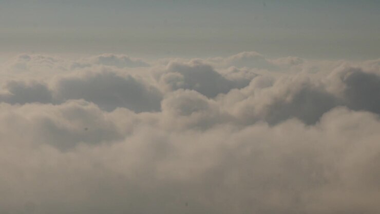 Clouds drift across a pale blue sky in a wide aerial view above a dense cloud layer. Soft daylight reveals textured cumulus formations and a calm high-altitude atmosphere