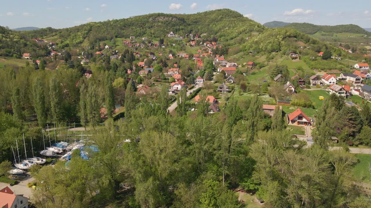 An aerial view of a small town nestled among lush green hills and dense forests. Scattered houses with red rooftops are surrounded by trees, creating a peaceful rural scene under a clear blue sky.