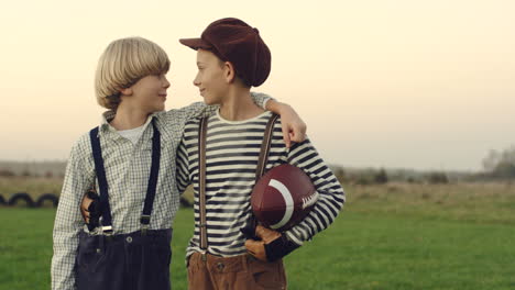 retrato de los dos adolescentes amigos jugadores de fútbol americano parados con una pelota y abrazándose en el campo del campo, mirándose con sonrisas y luego a la cámara