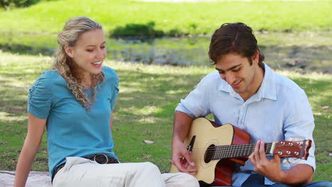 un hombre toca la guitarra y canta a una mujer mientras se sientan en el parque