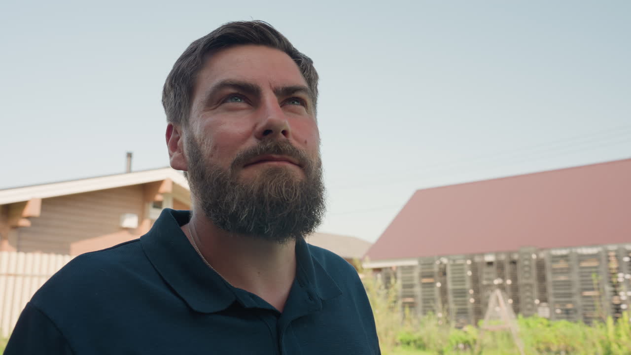 Bearded gardener in dark polo with tool apron stands beside blue drum with wooden crate, looking ahead in sunny backyard near house and barn