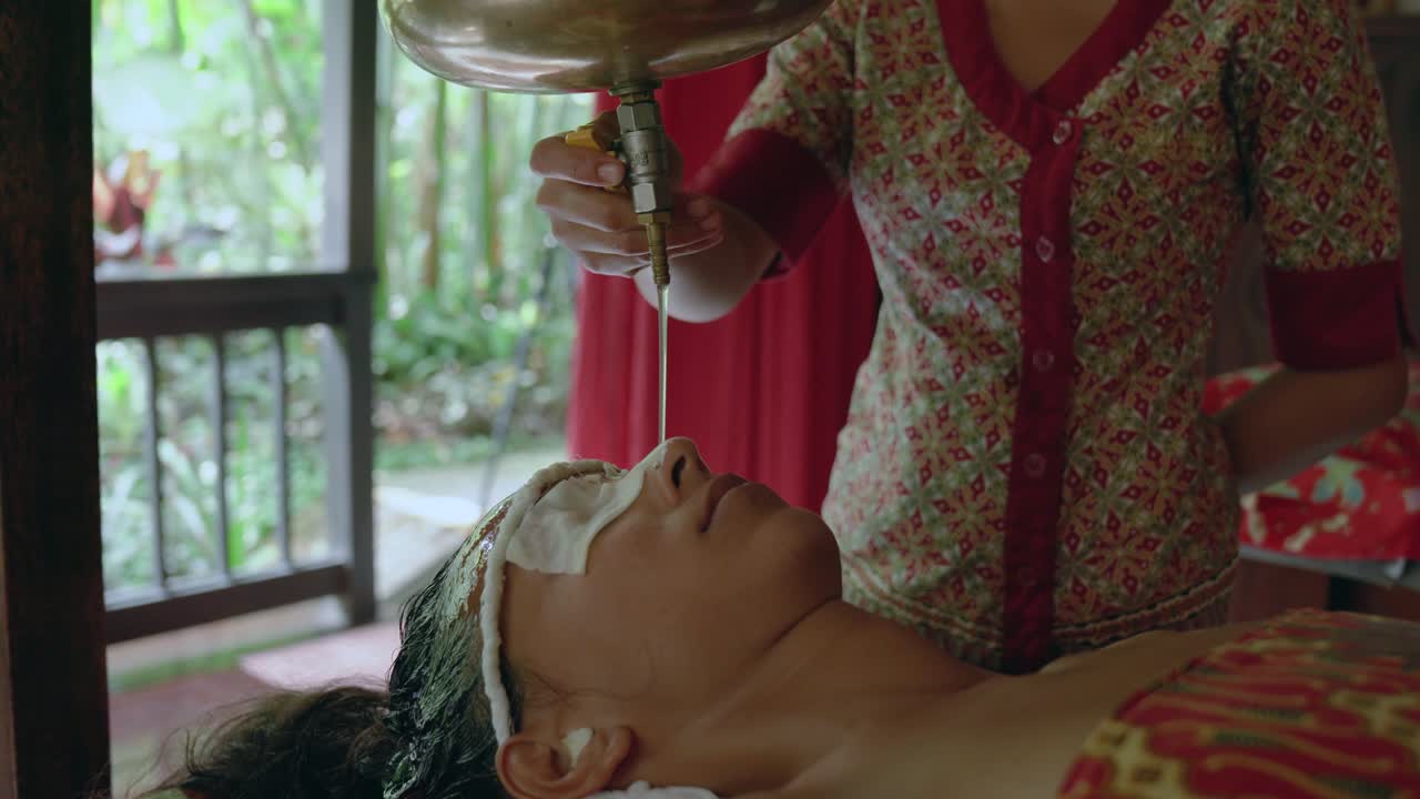 Nainen rentoutuu yksityisessä huoneessa vastaanottaessaan stressiä lievittävää Shirodhara-öljyhoitoa ayurvedisessä retriitissä
