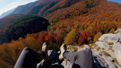 first-person perspective of legs dangling over a cliff edge with vibrant autumn mountains