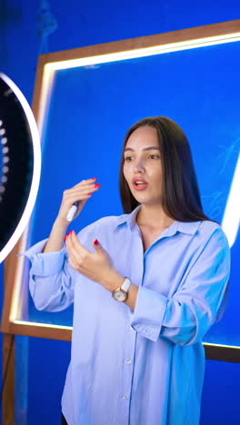 confident brunette with long hair talks to camera and gestures with her hands. camera on tripod and ring light is in front of lady. vertical video.